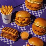 Flat lay of burgers, fries, and dipping sauce from Trip Burger restaurant arranged on a purple checkered surface with the slogan ‘Happiness in Every Bite.
