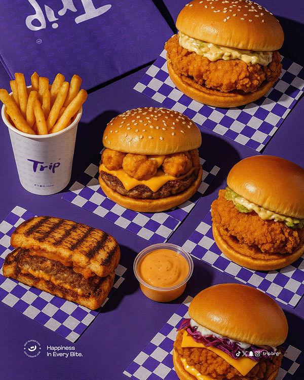 Flat lay of burgers, fries, and dipping sauce from Trip Burger restaurant arranged on a purple checkered surface with the slogan ‘Happiness in Every Bite.