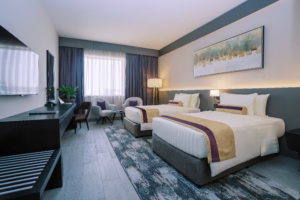 Twin hotel room with two single beds, lounge chairs, a floor lamp, and framed art, featuring a modern design with blue and beige tones.