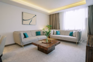 Spacious hotel room with a king-size bed, modern furniture, soft gray tones, and natural lighting from a curtained window.