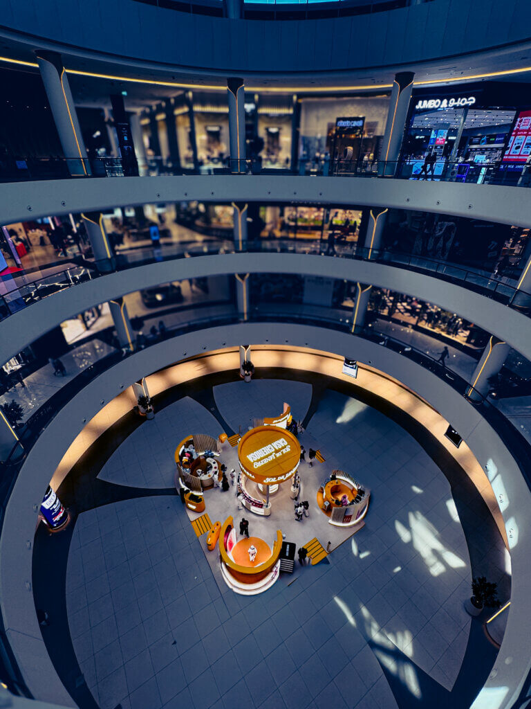 Modern shopping mall interior with circular multi-level design, central event space featuring promotional booths, and luxury retail stores in the background.