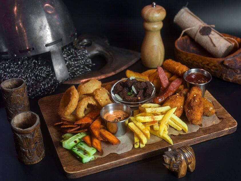 Rustic wooden platter with assorted fried snacks including onion rings, French fries, sweet potato fries, cucumber slices, chicken strips, sausages, beef bites, dipping sauces, and condiments, styled with medieval props