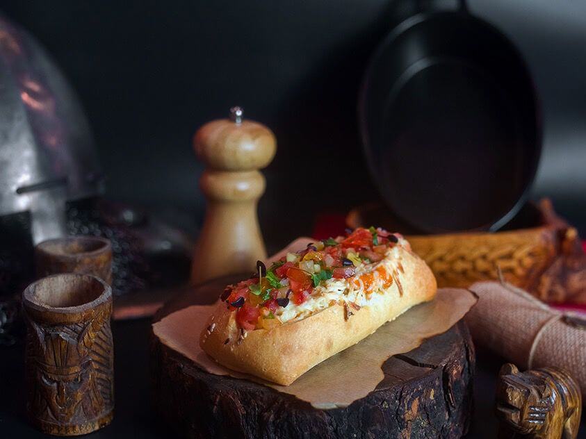 Creative portfolio showcase featuring a rustic food photography scene with a gourmet hotdog topped with colorful diced vegetables and cheese, styled on a wooden log surrounded by medieval props including wooden cups, a pepper grinder, and a Viking helmet.
