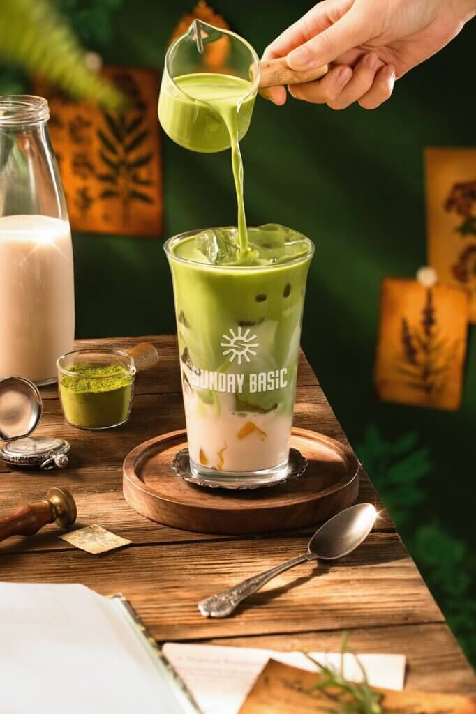 Creative portfolio showcase highlighting a close-up pour shot of creamy matcha over ice in a glass labeled Sunday Basic, styled with elegant accessories, wooden textures, and warm lighting for an artisanal café vibe.