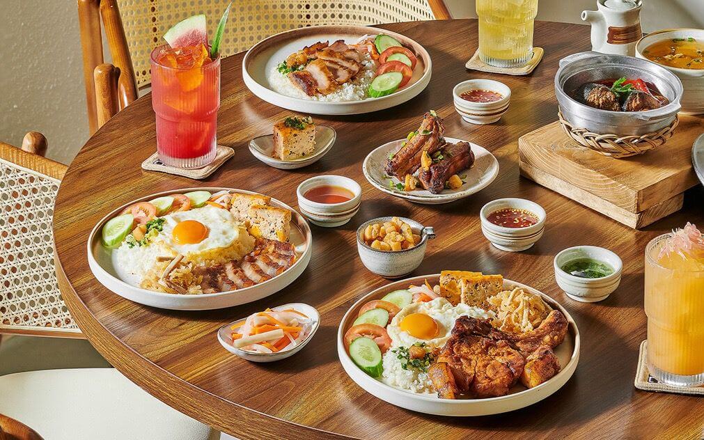 "Creative portfolio showcase of bright and inviting restaurant food photography showing a full Asian meal spread with rice, eggs, grilled meats, sauces, and fresh drinks on a wooden table under natural light.